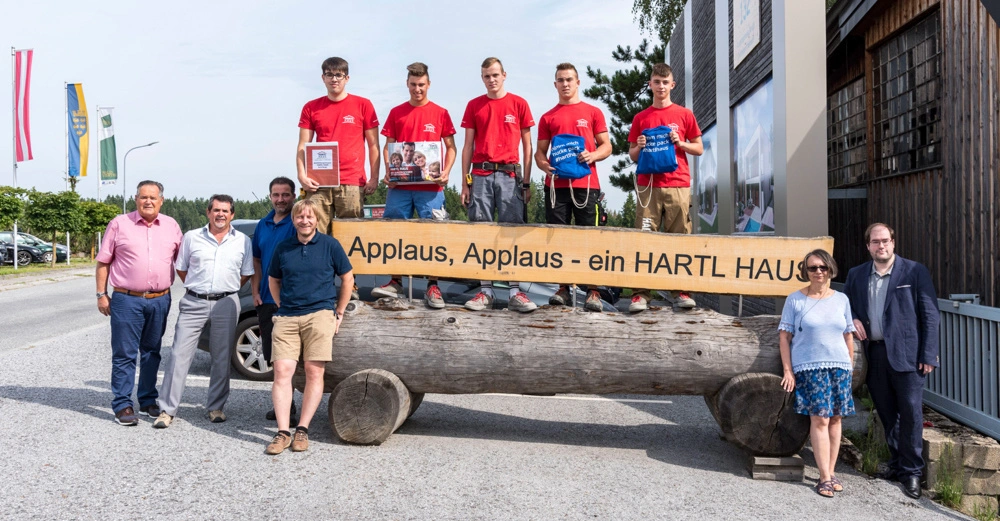 Eine Gruppe von Menschen posiert vor einem Holzstamm mit einem Schild Applaus Applaus - ein HARTL HAUS einige junge Männer in roten Hemden halten Preise sie stehen draußen auf einer Straße Flaggen im Hintergrund.