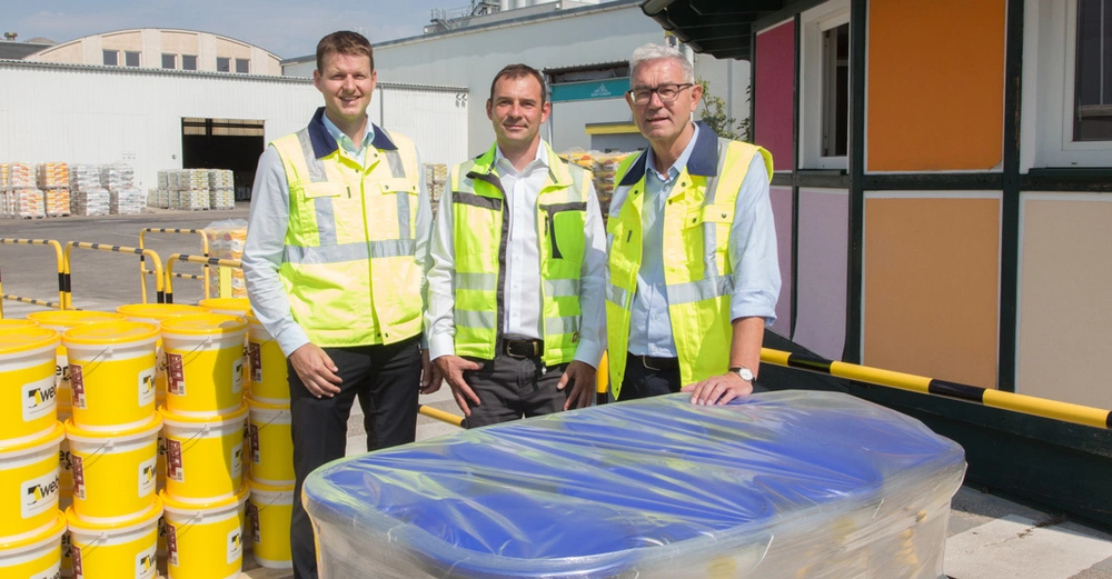 3 Vertreter aus dem Baumit-Management stehen in weißem Hemd, gelber Baustellenweste auf dem Firmengelände. Im Hintergrund ein Stapel gelbe Kisten, im Vordergrund eine Palette Dämmmaterial. 