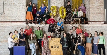 Eine Gruppe von etwa 25 Menschen posiert fröhlich mit goldenen Luftballons mit der Zahl 30 vor einer Backsteinwand. Konfetti fliegt herum. Auf Holzkiste steht AZW.