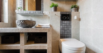 Eine moderne Toilette steht in einem Badezimmer mit beigen Fliesen. Links gibt es ein Waschbecken aus Stein auf einer steinernen Arbeitsplatte. Pflanzen und Handtücher sind dekorativ positioniert.