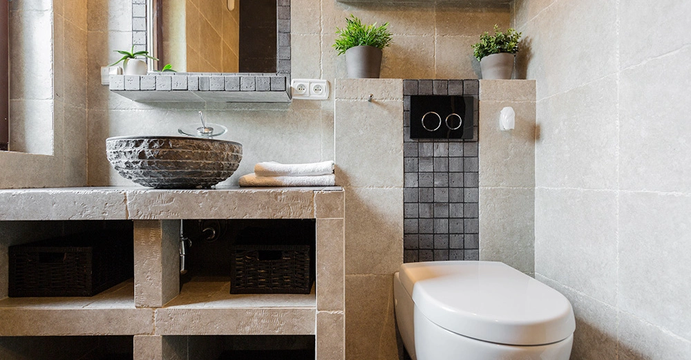 Eine moderne Toilette steht in einem Badezimmer mit beigen Fliesen. Links gibt es ein Waschbecken aus Stein auf einer steinernen Arbeitsplatte. Pflanzen und Handtücher sind dekorativ positioniert.