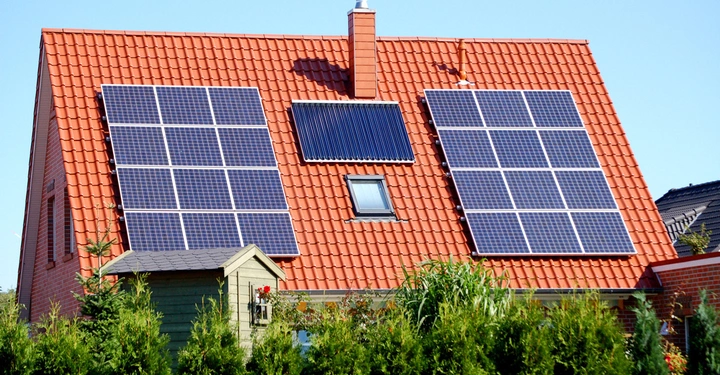 Solarpaneele und Sonnenkollektor auf dem Dach eines roten Ziegelhauses in einem grünen Garten bei sonnigem Wetter. Ein Schuppen steht vor dem Haus und Bäume umgeben es.
