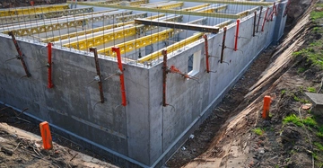 Betonfundament eines Gebäudes wird gebaut; Arbeiter benutzen Schalung und Stützen; Baustelle mit Erdarbeiten im Hintergrund.