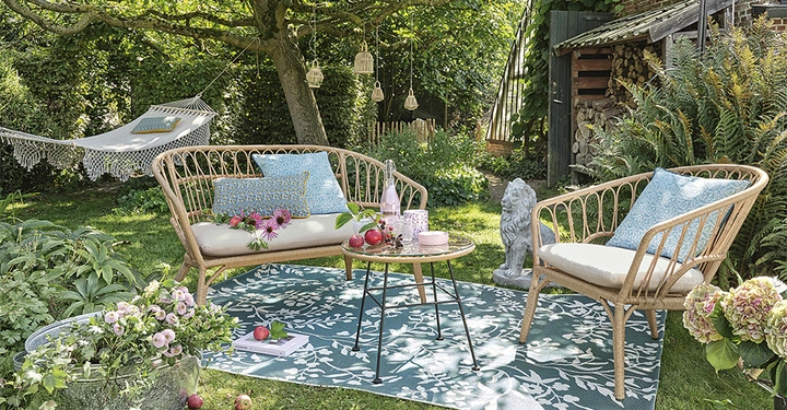 Galerie-Bild 6: Zwei geflochtene Sofas mit Kissen stehen auf einem blauen Teppich neben einem runden Tisch im Garten unter einem Baum Eine Hängematte hängt im Hintergrund umgeben von viel Grün und Blumen