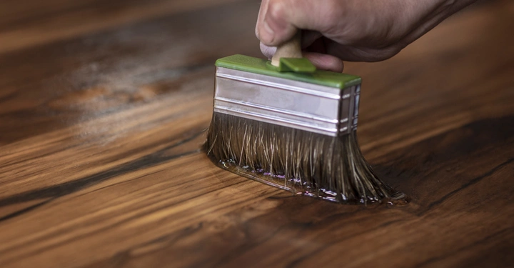 Eine Hand hält einen Pinsel und trägt Holzlack auf eine Holzfläche auf in einer Nahaufnahme.