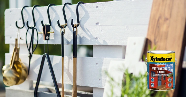 Dose Xyladecor Wetterschutz-Farbe steht vor einem weißen Holzzaun mit hängenden Gartengeräten im Freien