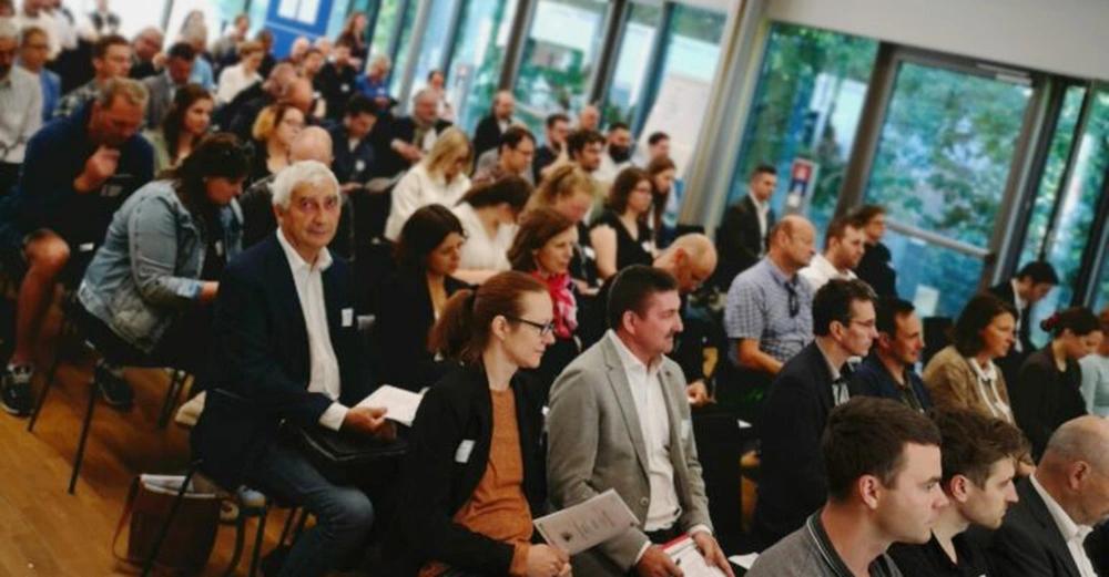 buntes Publikum hört interessiert einem Vortrag zu, im Hintergrund große Fensterflächen in Grün
