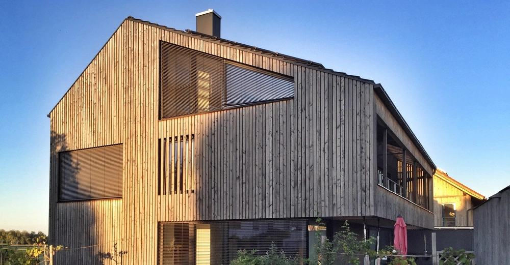 Gebäude bestehend aus Holzplanken ohne erkennbare Bewegung umgeben von einem Garten bei klarem Himmel und Sonnenuntergangslicht auf die Fassade fallend.