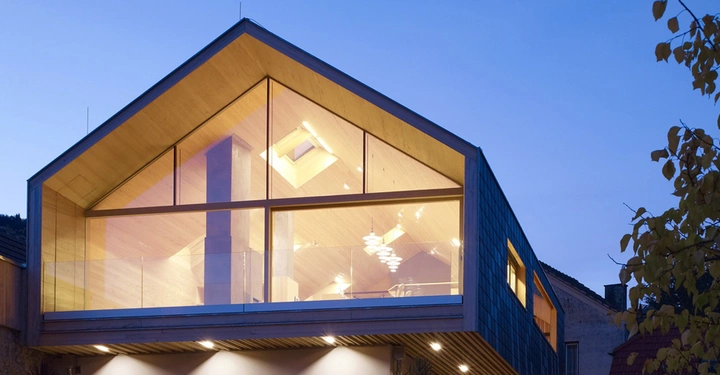 Haus mit geneigtem Dach leuchtet von innen in der Dämmerung eine aus Glas und Holz bestehende Fassade wird von modernen Leuchten erhellt umgeben von anderen Gebäuden und Bäumen