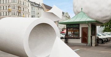 Eine übergroße Toilettenpapierrolle liegt aufgerollt auf einem Platz neben einem grünen Imbissstand mit der Aufschrift Würstelhütte umgeben von mehrstöckigen weißen Gebäuden mit verzierten Fassaden.
