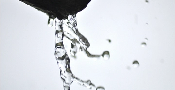 Nahaufnahme von klarem Wasser, das aus einem dunklen Rohr oder einer Kante fließt. Einzelne Tropfen lösen sich vom Wasserstrahl vor einem hellen, unscharfen Hintergrund.