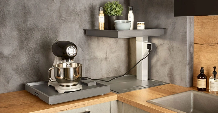 Küchenmaschine steht auf einer Arbeitsplatte in einer modernen Küche, daneben befindet sich eine erhobene Ablagefläche mit kleinen Gegenständen; im Hintergrund sind graue Wände und weitere Küchenutensilien zu sehen.