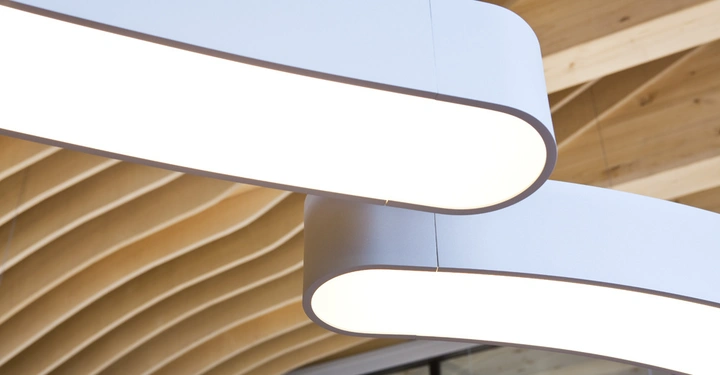 Leuchtendes Deckenlicht hängt von der Decke eines modernen Gebäudes. Im Hintergrund befinden sich wellenförmige Holzelemente und Glaswände, die eine offene und stilvolle Inneneinrichtung zeigen.