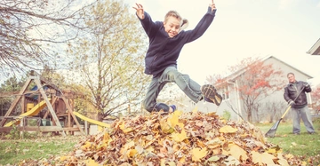 Kind springt in Laubhaufen im Garten während eine andere Person mit einem Rechen in der Nähe steht Spielplatz im Hintergrund Bäume und Häuser umgeben die Szene