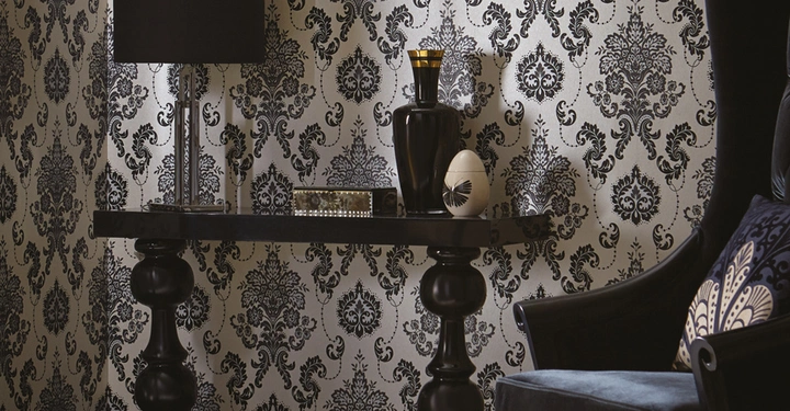 Galerie-Bild 7: Schwarze Tischlampe steht auf einem dekorativen schwarzen Tisch zusammen mit Vasen und Büchern in einem opulent dekorierten Raum mit gemusterten Tapeten und einem großen, gepolsterten Sessel.