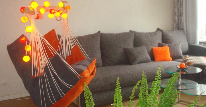 Hängesessel hängt beleuchtet von bunten Kugeln in einem Wohnzimmer mit grauem Sofa, orange Kissen und grünem Farn auf dem Boden. Fenster mit Vorhängen rechts im Hintergrund.