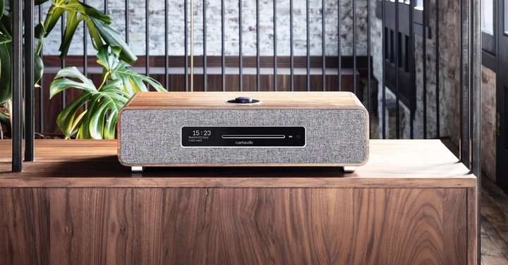 Ein graues Audiosystem steht auf einem Holztisch, es zeigt 15:23 auf einer Digitalanzeige. Ein Topfpflanze ist links dahinter. Raum im Hintergrund mit gemauerter Wand. Text: 15:23, Natalie (Radio mixes), 10:55, ruarkaudio