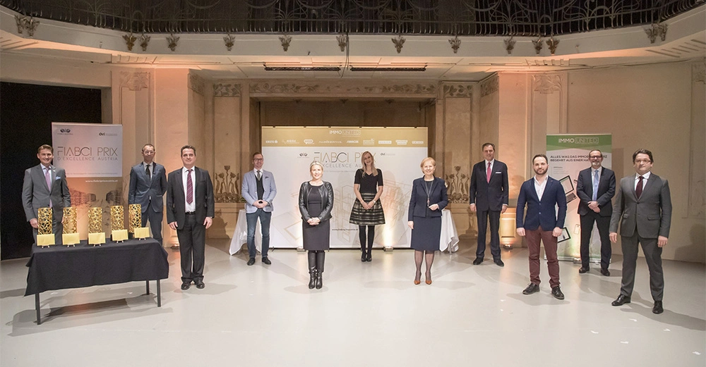 Elf Personen in formeller Kleidung stehen nebeneinander in einer großen Halle Im Hintergrund Banner mit der Aufschrift FIABCI PRIX D'EXCELLENCE AUSTRIA und IMMO UNITED Links auf einem Tisch fünf Trophäen.