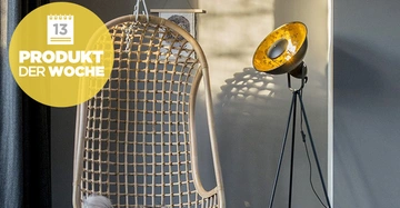 Ein hängender Rattanstuhl steht vor einem schattigen Vorhang neben einer eingeschalteten Stehlampe in modernem Raum. Gelber Kreis mit Kalender und Text: 13 PRODUKT DER WOCHE.