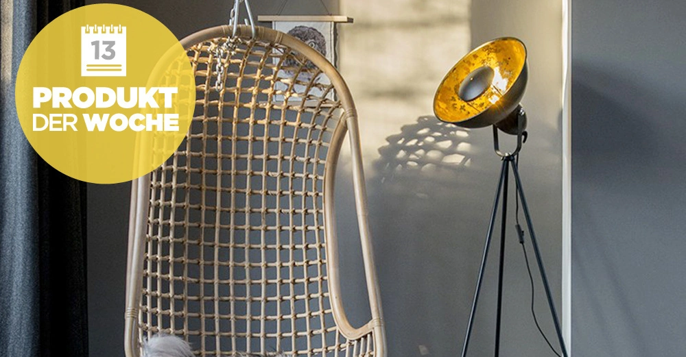 Ein hängender Rattanstuhl steht vor einem schattigen Vorhang neben einer eingeschalteten Stehlampe in modernem Raum. Gelber Kreis mit Kalender und Text: 13 PRODUKT DER WOCHE.
