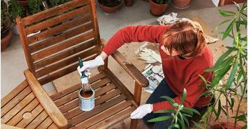 Frau hockt auf Terrasse und arbeiten mit Pinsel und Holzöl an Gartenstuhl aus Holz