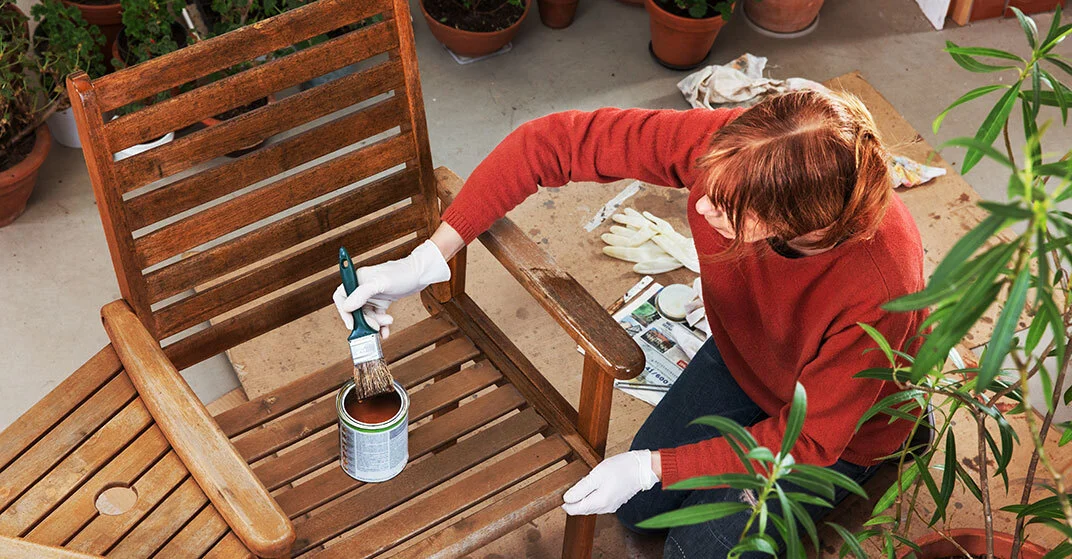 Frau hockt auf Terrasse und arbeiten mit Pinsel und Holzöl an Gartenstuhl aus Holz