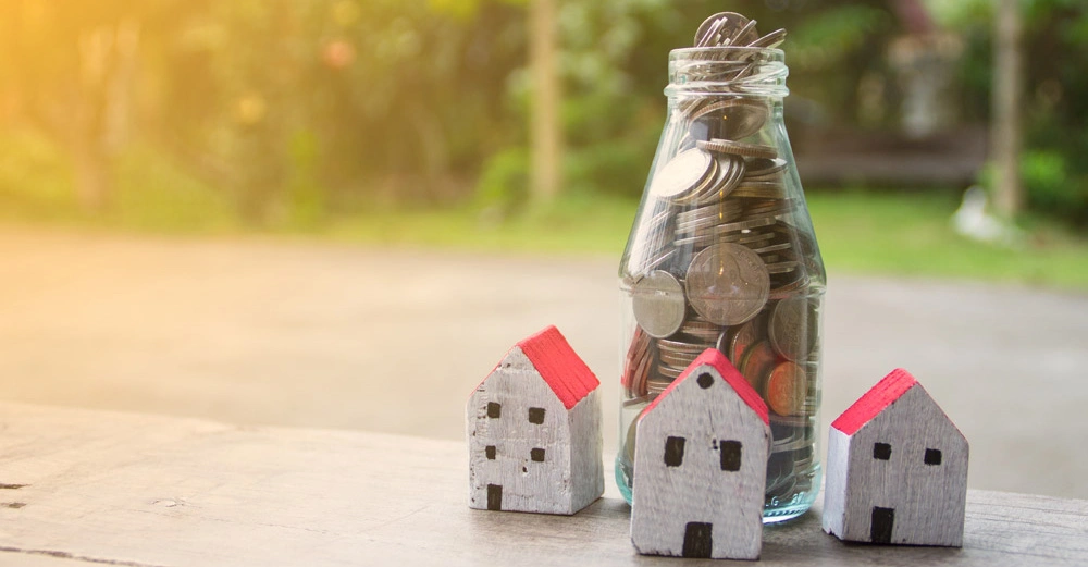 Drei kleine weiße Holzhäuser mit roten Dächern stehen neben einer Glasflasche voller Münzen auf einem Holztisch im Freien mit grünem Hintergrund.