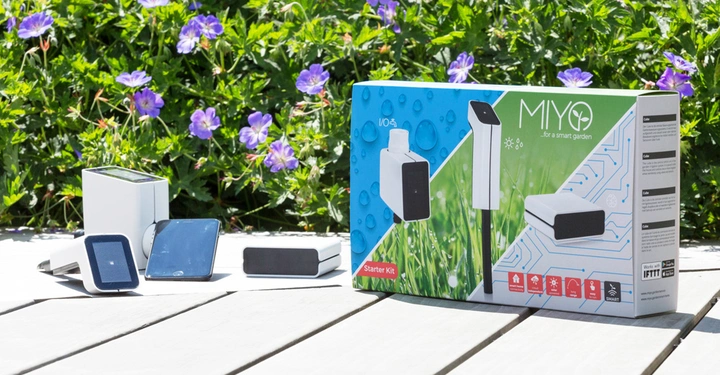 Ein Smart-Garten-Starter-Kit bestehend aus verschiedenen weißen Sensorgeräten liegt auf einem Holztisch im Freien vor einer grünen Pflanze mit lila Blüten; die Verpackung zeigt Produkte und Anleitungen. Text: MIYO for a smart garden Starter Kit.