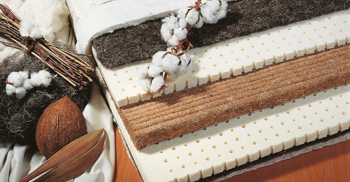 Galerie-Bild 4: Geschichtete Matratze mit unterschiedlichen Materialien liegt auf einem Holzboden. Daneben liegen Baumwollzweige, Kokosnuss, Stoff und Naturfasern, die die Verwendung natürlicher Materialien verdeutlichen.
