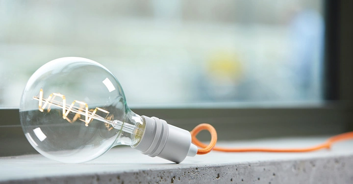 Galerie-Bild 4: Glühbirne liegt auf einer Betonoberfläche in der Nähe eines Fensters und ist mit einem orangefarbenen Kabel verbunden. Das Fenster bietet einen unscharfen Blick nach draußen.