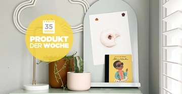 Gelbe Blase mit der Aufschrift "35 PRODUKT DER WOCHE" vor einer Lampe, zwei Topfpflanzen und einem weißen Brett mit einem Bild eines Donuts und einem Kinderbuch in einem Raum.