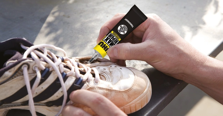 Eine Hand drückt Kleber aus einer Tube auf die Sohle eines weißen Sportschuhs mit schwarzen Streifen im Freien. Text auf der Tube UHU MAX REPAIR EXTREME.