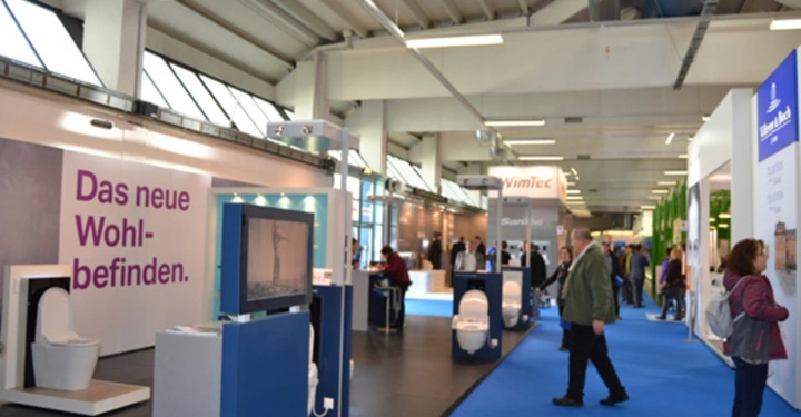 Galerie-Bild 5: Toiletten und Sanitäranlagen werden von Menschen auf einer Messe besichtigt. Die Wände sind mit Firmenlogos und Displays dekoriert. Text auf der linken Seite lautet Das neue Wohlbefinden.