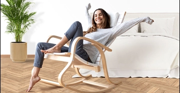 Eine lächelnde Frau sitzt entspannt in einem hellen Schlafzimmer auf einem geschwungenen Holz-Schaukelstuhl. Sie streckt die Arme aus, trägt ein graues Langarmshirt und dunkle Hose. Im Hintergrund stehen ein großes Bett mit heller Bettwäsche und eine grüne Zimmerpflanze auf einem Parkettboden.