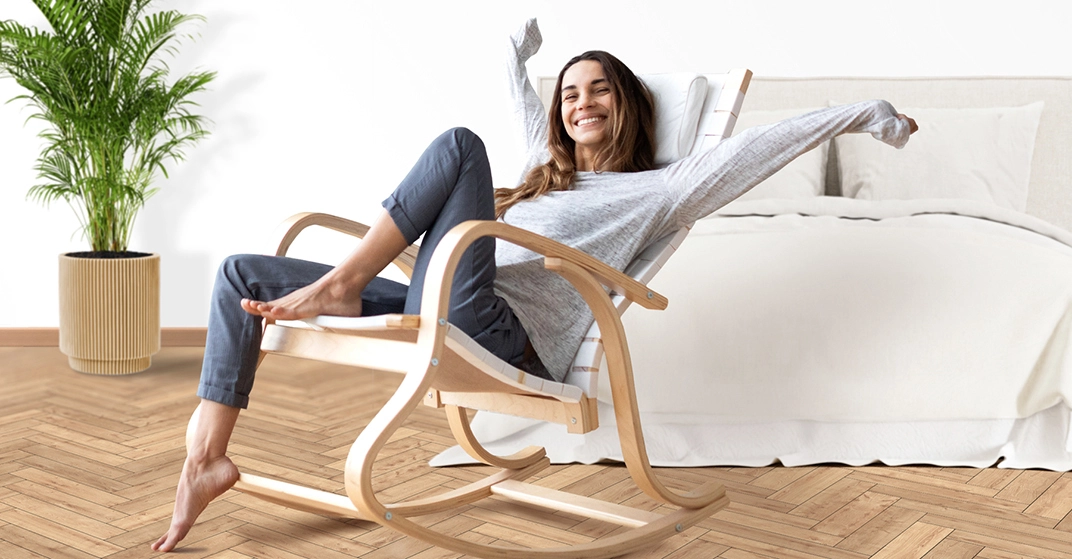 Eine lächelnde Frau sitzt entspannt in einem hellen Schlafzimmer auf einem geschwungenen Holz-Schaukelstuhl. Sie streckt die Arme aus, trägt ein graues Langarmshirt und dunkle Hose. Im Hintergrund stehen ein großes Bett mit heller Bettwäsche und eine grüne Zimmerpflanze auf einem Parkettboden.