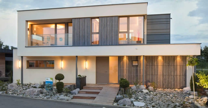 Modernes zweistöckiges Haus mit großen Fenstern und Balkon; beleuchtet von warmen Lichtern; umgeben von Steingarten und Gehweg in einer Vorortsiedlung.