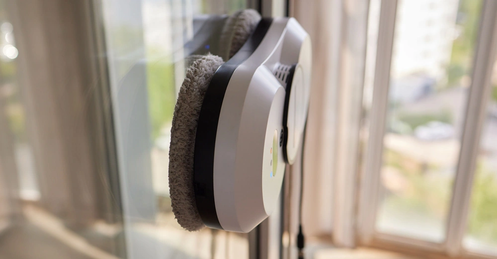 Ein automatischer Fensterreinigungsroboter reinigt eine große Glasfensterfläche. Das Gerät befindet sich in einem Wohnbereich mit verschwommenem Blick auf die Außenseite bei Tageslicht.