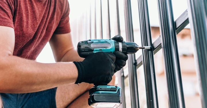 Eine Person in einem roten T-Shirt verwendet eine Bohrmaschine, um ein Metallgitter zu befestigen. Die Umgebung wirkt wie ein Außenbereich mit unscharfem Hintergrund.
