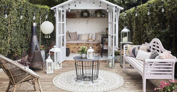 Gartenlaube mit Sofa und Kissen und Teelichtern auf Holzboden umgeben von Hecken mehrere Laternen und Lichterketten hängen überall ein Eisenofen und Sitzmöbel befinden sich im Außenbereich eines gemütlichen Gartenbereichs