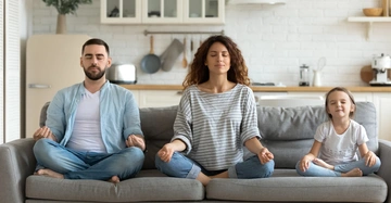Drei Personen sitzen im Schneidersitz auf einem Sofa meditierend in einem hellen Wohnzimmer mit Küche im Hintergrund