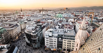 Verschieden hohe Gebäude und Kirchen mit grünen Kuppeln und spitzen Türmen stehen in der Innenstadt bei Sonnenuntergang umgeben von Menschenmengen und Kränen sowie Hügeln im Hintergrund.