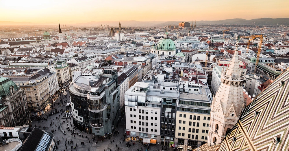 Verschieden hohe Gebäude und Kirchen mit grünen Kuppeln und spitzen Türmen stehen in der Innenstadt bei Sonnenuntergang umgeben von Menschenmengen und Kränen sowie Hügeln im Hintergrund.