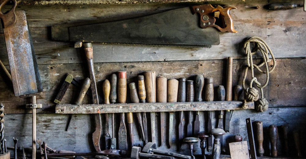 Werkzeuge hängen und stehen unbenutzt in einem Holzregal an einer rustikalen Holzwand in einer Werkstatt.