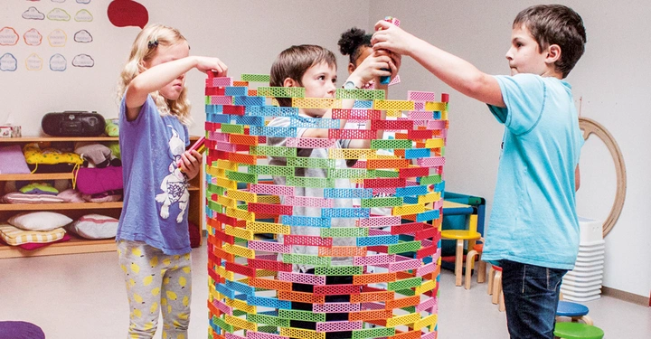 Kinder bauen mit bunten Bausteinen einen hohen Turm im Klassenzimmer Stehende Regale mit Kissen und Stühlen im Hintergrund Wände sind mit verschiedenen Zeichnungen und Papierdekorationen geschmückt