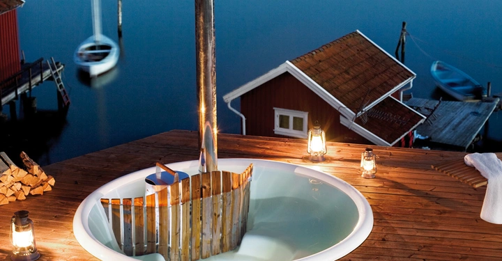 Galerie-Bild 3: Rundes Whirlpool steht in einem Holzdeck mit Laternen drumherum und Holzstapeln daneben mit Blick auf ein ruhiges Gewässer und ein rotes Gebäude am Ufer.
