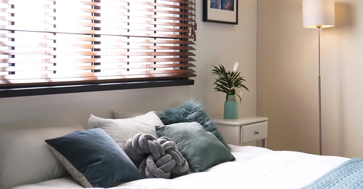 Schlafzimmer mit großem Doppelbett unter Fenster. Blaue und graue Zierkissen auf Bett, blaue Tagesdecke im unteren Rand. Fenster mit dunklen Holzjalousien innen versehen
