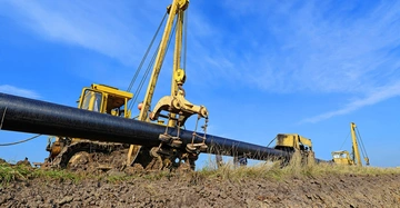 gelber Bagger steht an Baugrubenrand und hebt mit Greifarm schwarzes Abwasserrohr an, Hintergrund tiefblauer Himmel