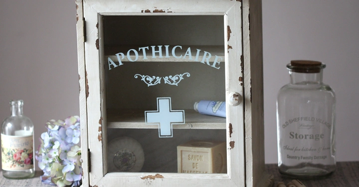 Galerie-Bild 13: Weißer Medizinschrank mit Glastür steht still auf einem Tisch umgeben von Glasflaschen und Seife; der Schrank trägt die Aufschrift APOTHECAIRE und ein Plus-Symbol.