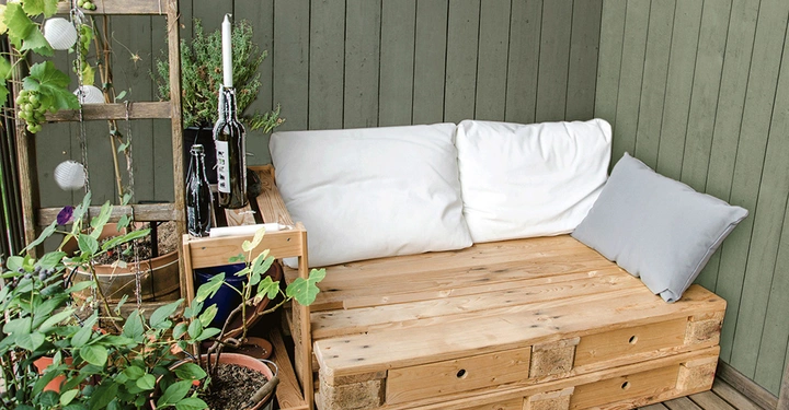 Holzsofa mit weißen Kissen steht in einer Ecke eines Balkons, links davon befinden sich Pflanzen in Töpfen und dekorative Weinflaschen mit Stabkerzen. Hintergrund aus grünen Holzwänden.