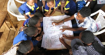Kinder in Schuluniformen sitzen um einen Tisch im Klassenzimmer und arbeiten gemeinsam an großen Papierbögen mit Skizzen und Notizen. Text ist nicht vorhanden